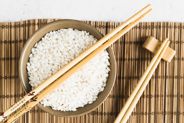 Palillos de madera y taza con arroz blanco en estera de bambú