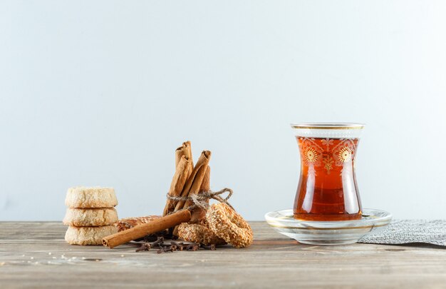 Palitos de canela con galletas, clavo de olor, un vaso de té, mantel vista lateral en madera y pared blanca