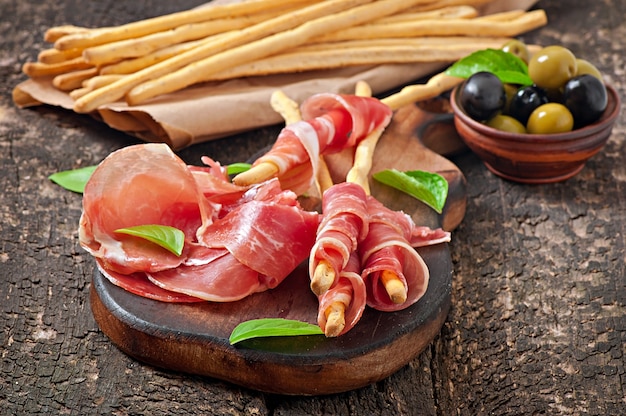 Palitos de pan grissini con jamón, aceitunas, albahaca sobre madera vieja