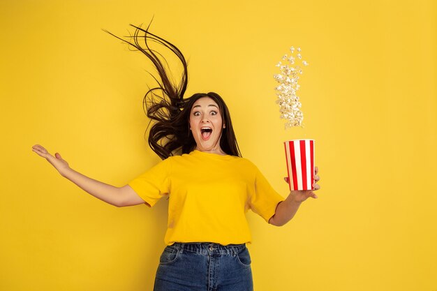 Palomitas de maíz voladoras y asombradas. Retrato de mujer caucásica aislado en pared amarilla. Hermosa modelo morena en casual. Concepto de emociones humanas, expresión facial, ventas, anuncios, copyspace.