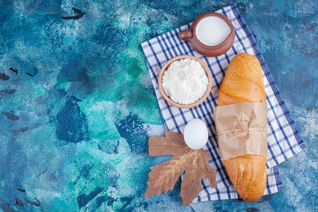 Pan baguette, harina, huevo y leche sobre un paño de cocina, sobre el fondo azul.