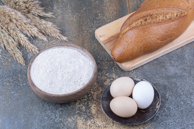 Pan de bastón en una placa junto a tallos de trigo, tazón de harina y huevos en la superficie de mármol