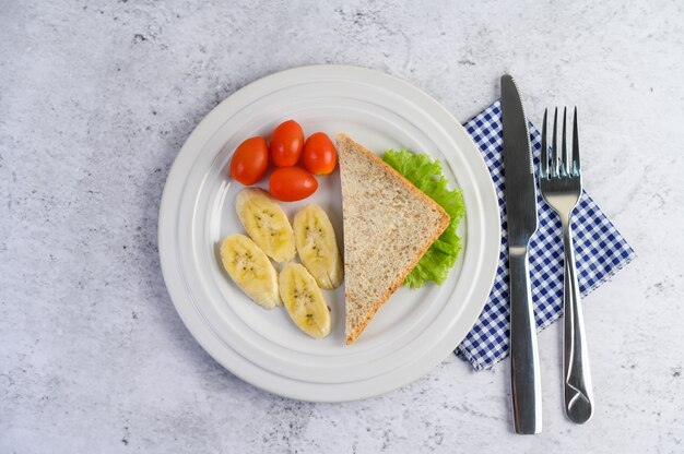 Pan, plátano y tomate en un plato blanco con tenedor y cuchillo.