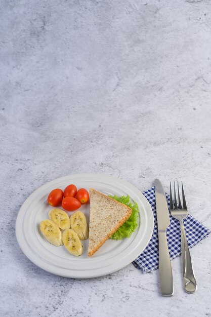 Pan, plátano y tomate en un plato blanco con tenedor y cuchillo.