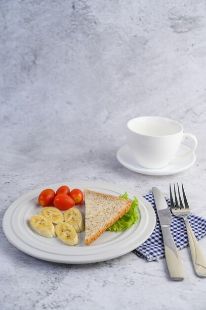 Pan, plátano y tomate en un plato blanco con tenedor y cuchillo.