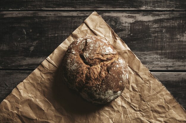 Pan redondo de grano entero de centeno saludable en papel artesanal marrón aislado en mesa de madera de granja negra.