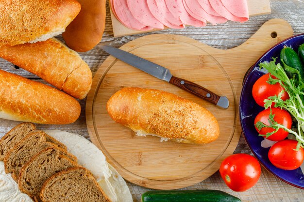 Pan con tomates, pepinos, cuchillo, salchichas, verduras planas en madera y tabla de cortar