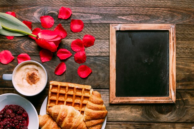 Panadería en un plato cerca de una taza de bebida, flor, mermelada, pétalos y marco de fotos