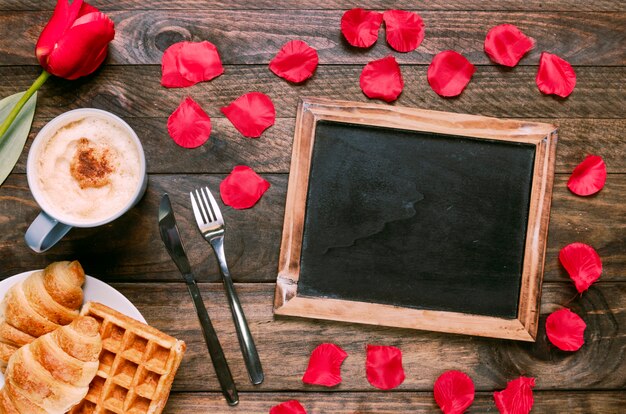 Panadería en un plato cerca de una taza de bebida, flores, cubiertos, pétalos y marco de fotos