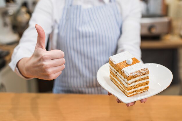 Panadero femenino que sostiene los pasteles frescos en la placa de cerámica de la forma del corazón que muestra el pulgar encima de la muestra