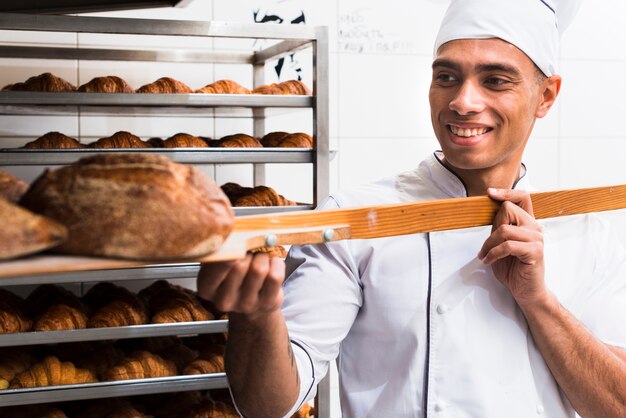 Panadero hombre sonriente en uniforme sacando con pala pan recién horneado del horno