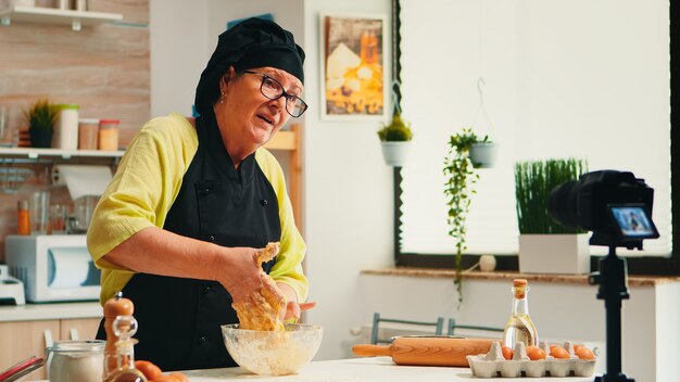 Panadero maduro con experiencia grabando un tutorial de recetas culinarias con una cámara moderna en la cocina con bonete y delantal. Chef influyente que utiliza la tecnología de Internet comunicándose en las redes sociales.