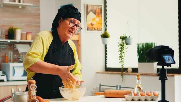 Panadero maduro con experiencia grabando un tutorial de recetas culinarias con una cámara moderna en la cocina con bonete y delantal. Chef influyente que utiliza la tecnología de Internet comunicándose en las redes sociales.