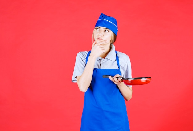 Foto gratuita panadero mujer en delantal azul sosteniendo una sartén tefal y parece confundida y pensativa