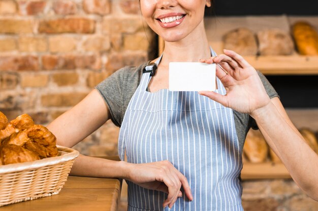 Panadero de sexo femenino joven sonriente que muestra la tarjeta de visita blanca en la tienda de la panadería