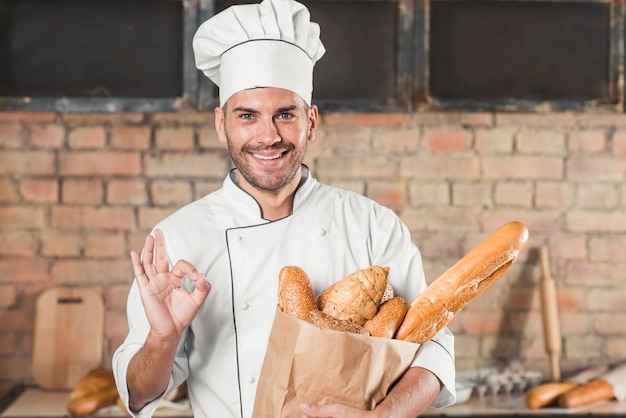 Panadero de sexo masculino joven sonriente que muestra la muestra aceptable que sostiene la barra de pan