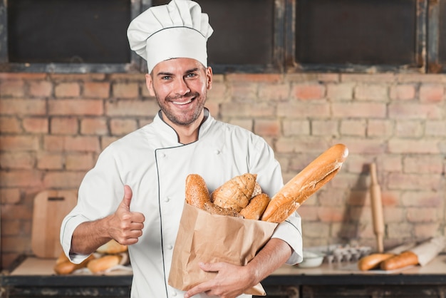 Panadero de sexo masculino joven sonriente que sostiene el pan de panes en bolsa de papel que muestra el pulgar encima de muestra