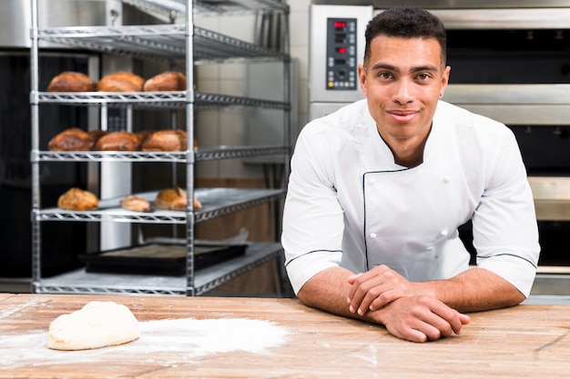 Panadero de sexo masculino de pie detrás de la mesa con masa en la panadería