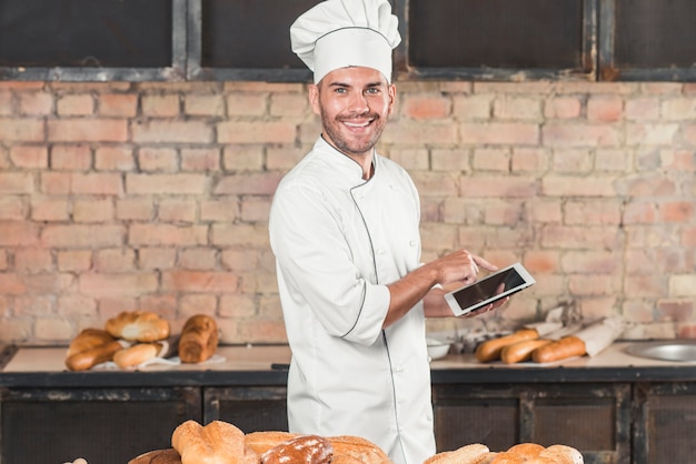 Panadero de sexo masculino sonriente que usa la tabla digital que se coloca detrás de la tabla con panes cocidos