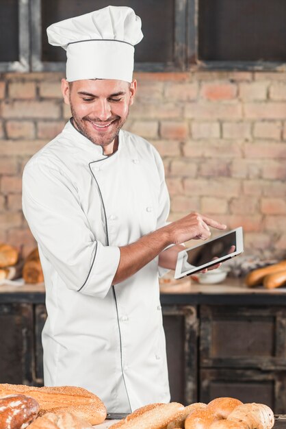 Panadero de sexo masculino sonriente que usa la tableta digital que mira panes cocidos en la tabla