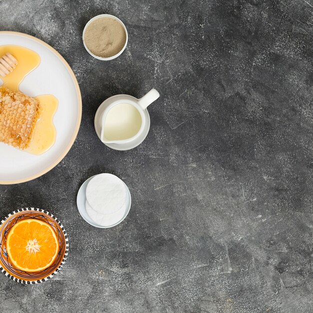 Panal sobre plato de cerámica con naranja a la mitad; almohadillas de algodón; jarra de leche y arcilla rhassoul sobre fondo de hormigón negro