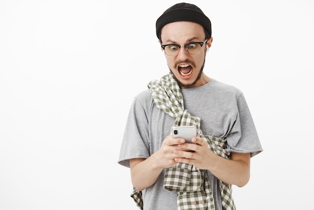 en pánico, chico sorprendido enviando una selección desnuda a mamá por error gritando y mirando tembloroso a la pantalla del teléfono inteligente con auriculares inalámbricos y gorro negro sobre una pared blanca