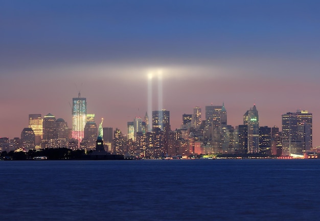 Panorama del horizonte del centro de Manhattan de Nueva York por la noche con la estatua de la libertad y haces de luz en memoria del 11 de septiembre visto desde el paseo marítimo de Nueva Jersey.