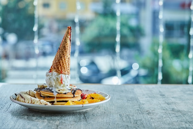 Foto gratuita panqueques con cono de helado, mandarina, fresa, plátano y jarabe de chocolate en el plato blanco sobre la superficie de mármol