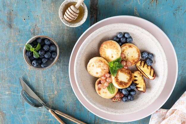 Panqueques de requesón, pasteles de queso, buñuelos de ricotta con arándanos frescos, grosellas y melocotones en un plato Desayuno saludable y delicioso para las vacaciones Fondo de madera azul