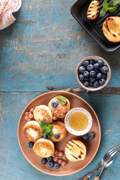 Panqueques de requesón, pasteles de queso, buñuelos de ricotta con arándanos frescos, grosellas y melocotones en un plato Desayuno saludable y delicioso para las vacaciones Fondo de madera azul