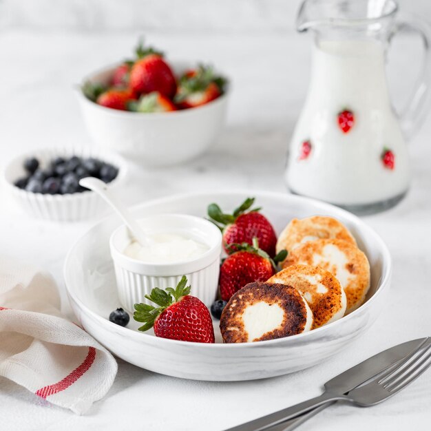 Panqueques de requesón, pasteles de queso, buñuelos de ricotta con fresas frescas y arándanos de un plato de cerámica blanca Desayuno saludable y delicioso para las vacaciones