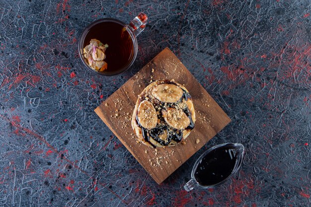 Panqueques con rodajas de plátano y vaso de té caliente sobre una superficie oscura.