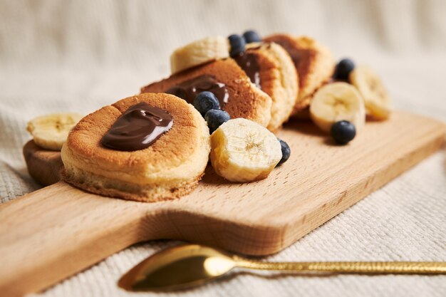 Panqueques con salsa de chocolate, bayas y plátano en una placa de madera detrás de un fondo blanco.