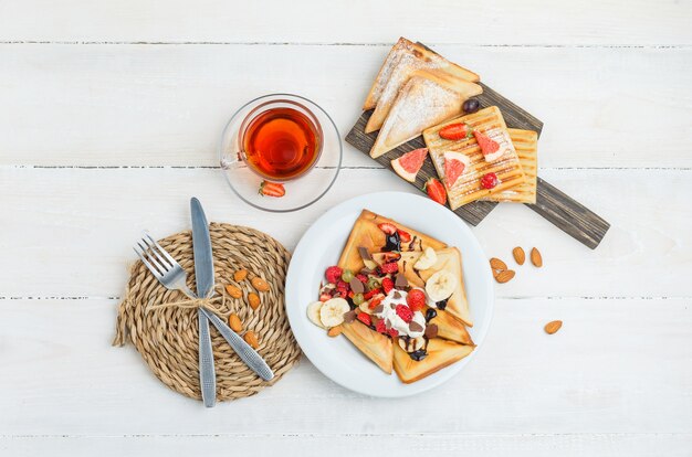 Panqueques sobre tabla de madera con té, almendras, cuchillo, tenedor, uvas y frambuesas