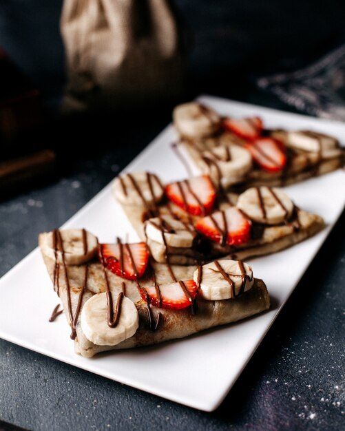 Panqueques de vista frontal en rodajas con fresas y plátanos dentro de un plato blanco sobre el escritorio gris