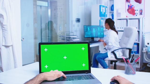 Pantalla de portátil pov con maqueta verde en gabinete de hospital. Médico que abre la puerta de cristal de la clínica. Medic usando un cuaderno con chroma key en exhibición en una clínica médica.