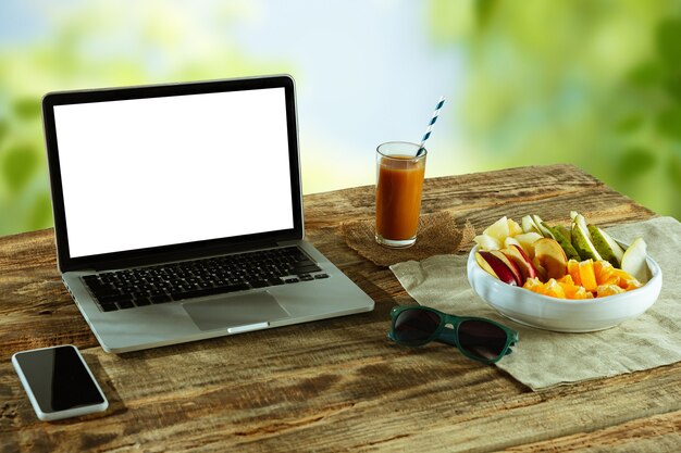 Pantallas en blanco de la computadora portátil y el teléfono inteligente en una mesa de madera al aire libre con la naturaleza en la pared Frutas y jugo fresco cercano. Concepto de lugar de trabajo creativo, negocio, autónomo. Copyspace.