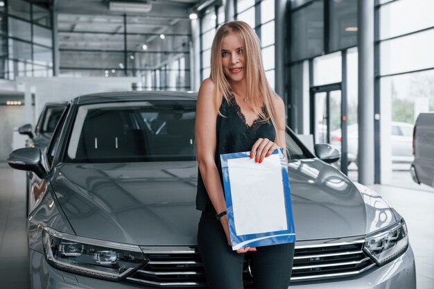 En pantalones negros. Chica y coche moderno en el salón. Durante el día en interiores. Comprar vehículo nuevo