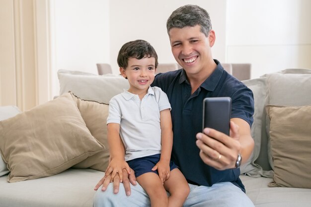 Papá alegre e hijo pequeño disfrutando del tiempo libre juntos, sentados en el sofá en casa, riendo y tomando selfie.
