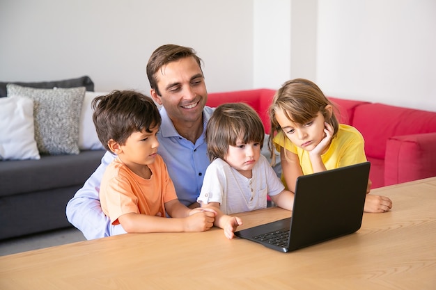 Papá alegre y niños pensativos viendo películas a través de la computadora portátil juntos durante el fin de semana. Padre feliz sentado a la mesa con los niños en la sala de estar. Concepto de paternidad, infancia y tecnología digital.