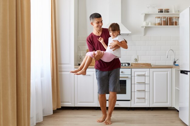 Papá alegre que lleva a su hija de cabello oscuro, jugando con un niño preescolar feliz en el interior contra el juego de cocina, una niña juguetona que se divierte con un padre sonriente en casa.