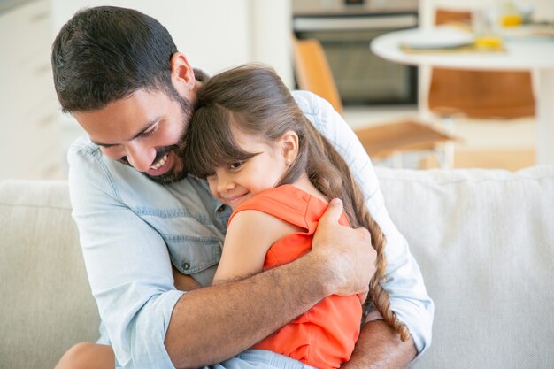 Papá alegre sentado con su niña en el sofá, abrazándola y abrazándola.