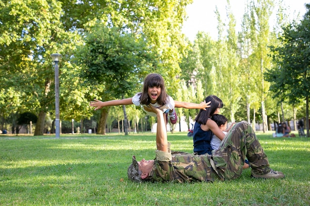 Papá amoroso tirado en la hierba y sosteniendo a la niña en manos rectas. Padre feliz en uniforme militar jugando con hija alegre. Mamá y niño sentados cerca de ellos. Concepto de reunión familiar y fin de semana.