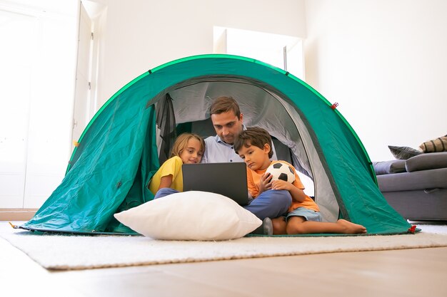 Papá cariñoso sentado con las piernas cruzadas con los niños en la carpa en casa y mirando la pantalla del portátil. Adorables niños viendo películas en la computadora con el padre. Concepto de infancia, tiempo en familia y fin de semana.