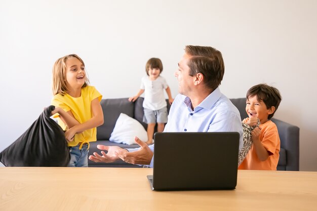 Papá caucásico hablando con niños juguetones y sentado en la mesa. Feliz padre de mediana edad con ordenador portátil cuando los niños juegan con la almohada en casa. Concepto de infancia y tecnología digital.