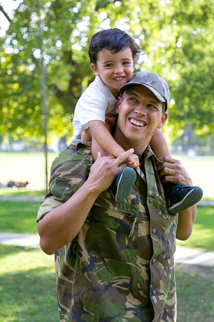 Papá caucásico con hijo en el cuello y sonriendo. Niño lindo feliz abrazando a padre en uniforme militar. Adorable niño caminando con papá en el parque de la ciudad. Concepto de reunión familiar, paternidad y regreso a casa