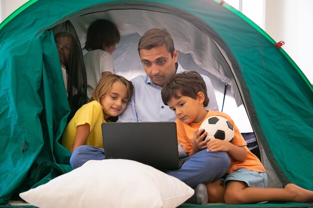 Papá caucásico sentado con las piernas cruzadas con los niños en la carpa en casa y viendo películas a través de la computadora portátil. Niños encantadores abrazando al padre, divirtiéndose y jugando. Concepto de infancia, tiempo en familia y fin de semana.