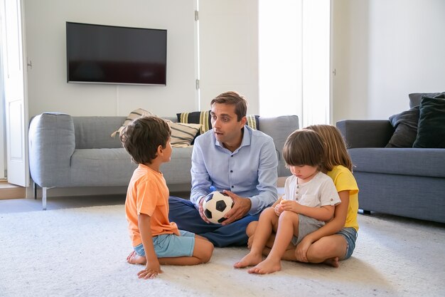 Papá caucásico sosteniendo la pelota y hablando con los niños. Amoroso padre e hijos de mediana edad sentados en el suelo en la sala de estar y jugar juntos. Concepto de infancia, actividad de juego y paternidad.