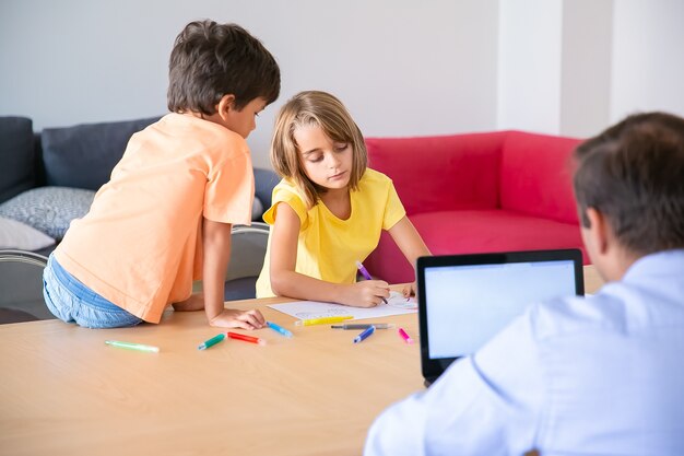 Papá caucásico trabajando en una computadora portátil y niños lindos pintando garabatos en la mesa. Dibujo de chica rubia concentrada con marcador y hermano mirándola. Concepto de infancia, creatividad y fin de semana.