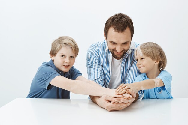 Papá e hijos no solo familia sino equipo. Retrato de feliz guapo hermanos y padre tomados de la mano mientras está sentado a la mesa, sonriendo ampliamente